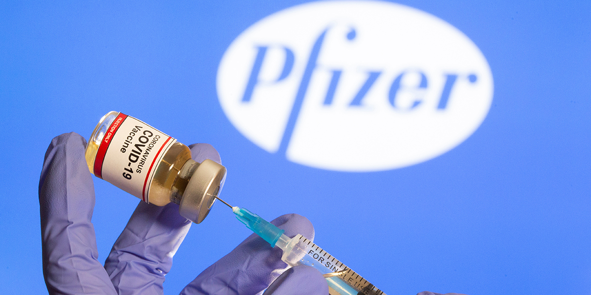 A woman holds a small bottle labeled with a "coronavirus covid 19 vaccine" sticker and a medical syringe in front of displayed pfizer logo in this illustration
